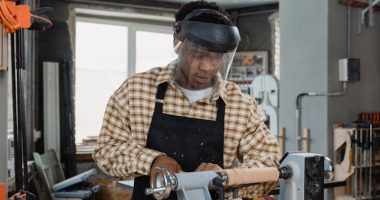 Man Wearing Checkered Long Sleeves and an Apron in a Workshop working on a lathe