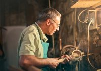 A senior craftsman focuses intently while working with tools in a dimly lit workshop.